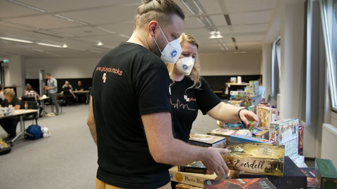Das Bild zeigt eine blonde Frau und einen Mann mit Zopf. Sie tragen FFP2-Masken und stehen vor einem Tisch, auf dem verschiedene Brettspiele liegen.