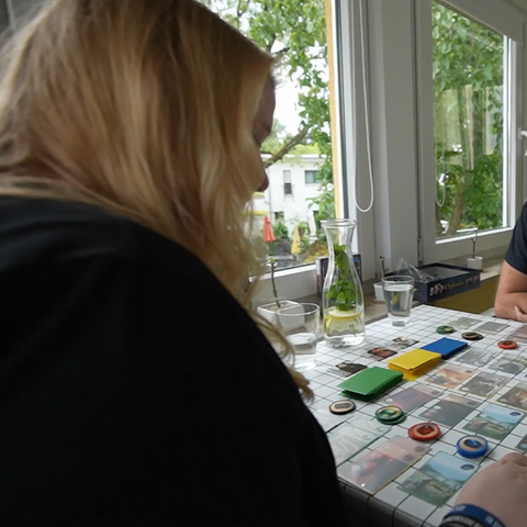 Das Bild zeigt eine blonde Frau und einen Mann mit Zopf. Sie sitzen an einem Holztisch an einem Fenster. Auf dem Tisch sind Spielkarten und -chips ausgebreitet.