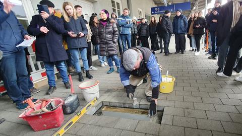 Ein Mann lässt auf dem Boden eines Bahnhofs eine Bronzeplatte ein, um ihn herum stehen Menschen.