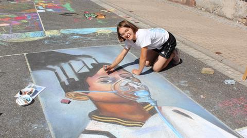 Ein Mädchen malt ein Bild mit einem Kind mit Gasmaske und einer Fabrik auf die Straße.