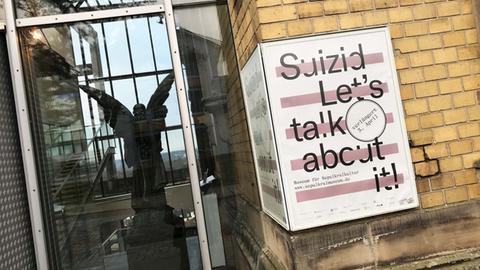 Foto mit einem Plakat, auf dem "Suizid - Let's talk about it" steht. Es hängt an der Hauswand neben dem Eingang zum Sepukralmuseum. Hinter dem gläserenen Eingang ist eine Engels-Statue im dunklen Inneren des Museums zu sehen.