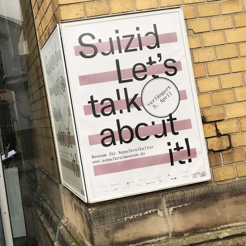 Foto mit einem Plakat, auf dem "Suizid - Let's talk about it" steht. Es hängt an der Hauswand neben dem Eingang zum Sepukralmuseum. Hinter dem gläserenen Eingang ist eine Engels-Statue im dunklen Inneren des Museums zu sehen.