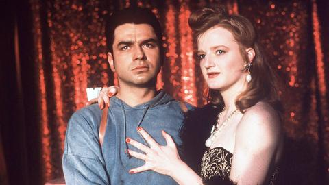 Das Szenenbild aus dem Film "Happy Birthday, Türke" zeigt die Schauspieler Hansa Czypionka und Nina Petri. Sie legt ihren Arm um ihn, er hebt ihren Oberschenkel hoch. Im Hintergrund ist ein glitzernder roter Vorhang zu sehen.