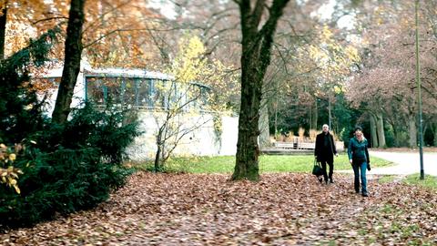 Filmstill des Drehortes "Pavilloncafé". Zwei Menschen spazieren an einem Gebäude im Park vorbei.