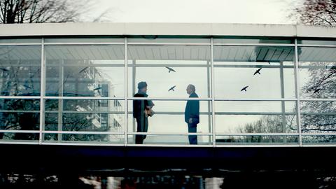 Filmstill des Drehortes "LKA-Gebäude". Zwei Männer stehen in einem verglasten Übergang.