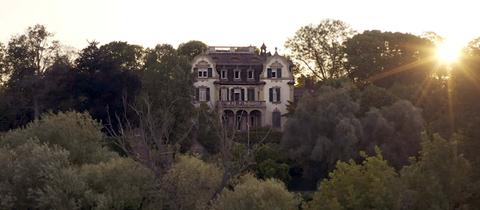 Filmstill des Drehortes "Villa". Eine historische Villa aus der fernen Vogelperspektive fotografiert, umgeben von vielen alten Bäumen.