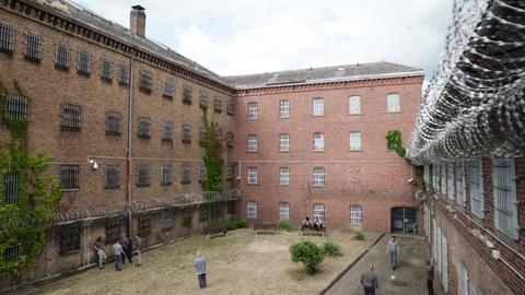 Filmstill des Drehortes "JVA". Blick in den Innenhof eines Gefängnisses, in welchem sich einige Häftlinge in blau-grauer Kleidung aufhalten.