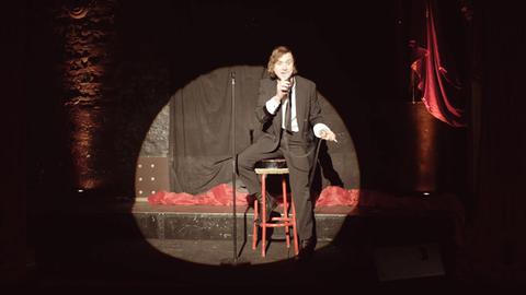 Eine dunkel abgehangene Bühne mit roten Vorhängen. Ein Spot beleuchtet einen Mann im schwarzen Anzug, der auf einem roten Barhocker sitzt und ein Mikrofon in der Hand hält.