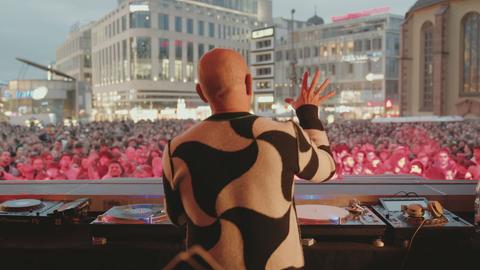 Das Bild zeigt einen Mann mit Glatze von hinten. Er trägt einen schwarz-weiß gemusterten Strickpullover und steht vor einem DJ-Pult. Vor ihm sind tanzende Menschen zu sehen.