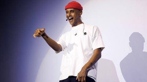 Ein dunkelhäutiger Mann mit rotem Beanie und weißem T-Shirt gestikuliert auf einer Bühne.