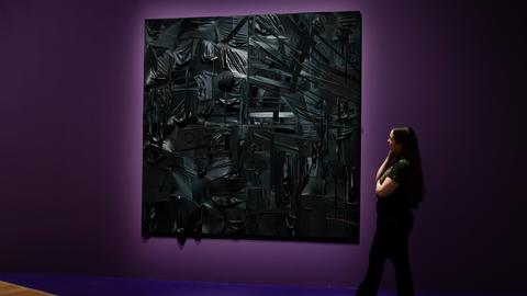 Das Bild zeigt eine in einem dunklen Lilaton gestrichene Wand mit einem quadratischen Kunstwerk aus schwarzen Kopfbedeckungen. Rechts am Bildrand steht eine schwarz gekleidete Frau mit langen dunklen Haare und betrachtet das Bild.