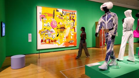 Das Bild zeigt eine junge Frau, die durch eine Ausstellungshalle läuft. An einer grün gestrichenen Wand hängen bunte Gemälde. Rechts am Bildrand sind Schaufensterpuppen aufgebaut, die Jogginganzüge tragen.