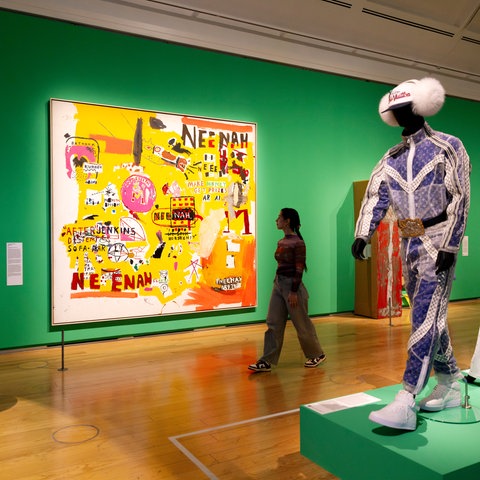 Das Bild zeigt eine junge Frau, die durch eine Ausstellungshalle läuft. An einer grün gestrichenen Wand hängen bunte Gemälde. Rechts am Bildrand sind Schaufensterpuppen aufgebaut, die Jogginganzüge tragen.