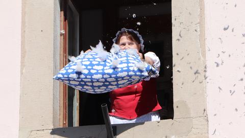 Eine Frau mit Kopfhaube schüttelt ein Kissen aus dem Fenster, Federn fliegen davon