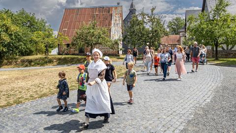 Eine Frau in ländlicher Tracht geht über eine gepflasterte Straße, gefolgt von Kindern und Erwachsenen in moderner Kleidung.