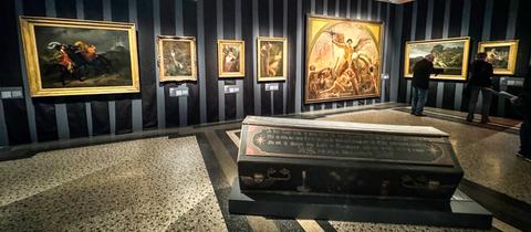 Ausstellungsansicht "Tod und Teufel" im Hessischen Landesmuseum Darmstadt. Das Bild zeigt einen dunklen Holzsarg in einem Eingangsbereich. Dahinter sind mehrere gerahmte Gemälde an einer schwarzen Wand angebracht.