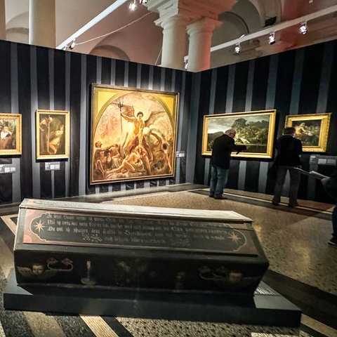 Ausstellungsansicht "Tod und Teufel" im Hessischen Landesmuseum Darmstadt. Das Bild zeigt einen dunklen Holzsarg in einem Eingangsbereich. Dahinter sind mehrere gerahmte Gemälde an einer schwarzen Wand angebracht.