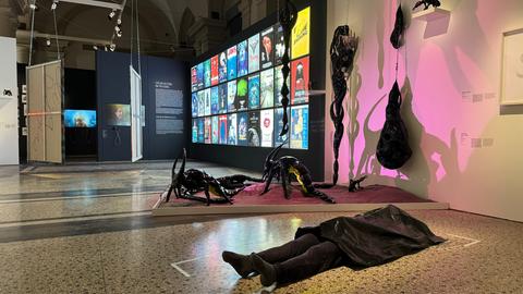 Ausstellungsansicht "Tod und Teufel" im Hessischen Landesmuseum Darmstadt. Zu sehen sind zwei spinnenartige Figuren, die auf einem grau gemusterten Steinboden liegen. Davor liegt eine in schwarz gekleidete Puppe mit einem schwarzen Tuch über dem Oberkörper und Gesicht. Im Hintergrund sind Filmposter an einer schwarzen Wand zu sehen.