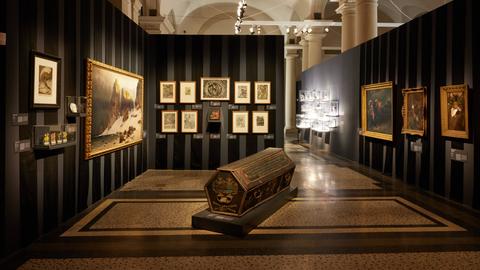 Das Bild zeigt einen Ausstellungsraum im Hessischen Landesmuseum in Darmstadt. An schwarzen Stellwänden hängen gerahmte Gemälde, in der Mitte steht ein Holzsarg auf einem Fliesenboden.