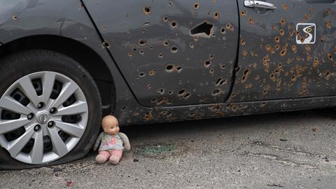 Das Bild zeigt ein silbernes Auto mit Einschusslöchern. Der rechte Hinterreifen ist platt. Am Reifen lehnt eine Puppe.