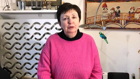 Eine Frau mit kurzen dunklen Haaren und pinkfarbenem Pullover sitzt an einem Esstisch. Hinter ihr hängen mehrere Gemälde an der Wand.