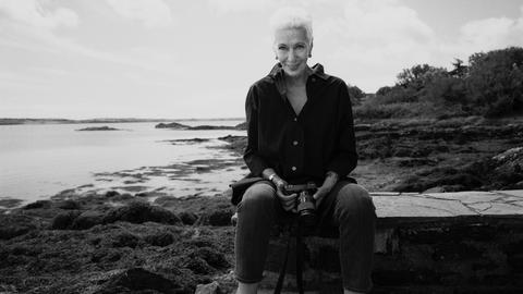 Das Schwarz-weiß-Bild zeigt eine Frau mit kurzen hellen Haaren, die auf einer Mauer sitzt. Sie hält eine Kamera. Im Hintergrund ist das Meer zu sehen.