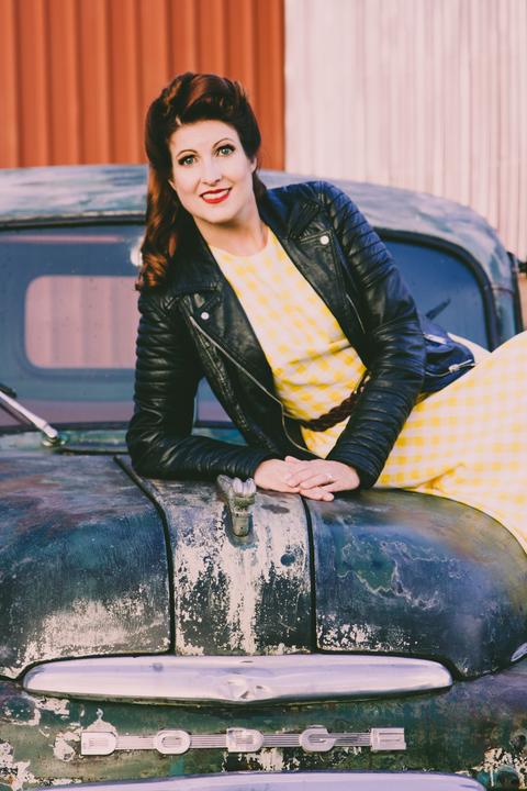Vanessa Hellmann-Koch, antoupiertes Haar, gelbes Kleid, Lederjacke - auf einen Oldtimer-Wagen gelehnt.