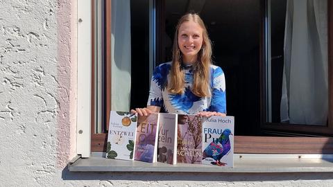 Eine Frau lehnt sich aus einem Fenster, vor ihr auf dem Fensterbrett stehen mehrere Bücher. SIe lacht in die Kamera.