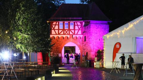 Das Bild zeigt einen mit Kopfsteinpflaster verlegten Hof, in dem Festzeltgarnituren und ein Pavillon aufgebaut sind. Im Hintergrund ist ein lila angeleuchtetes Torhaus im Fachwerkstil zu sehen. 