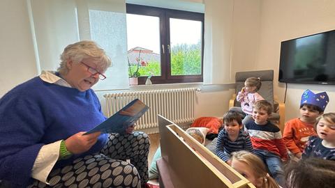 Das Bild zeigt eine ältere Frau mit kurzen grauen Haarren und blauem Pullover. Sie liest aus dem Buch "Der Regenbogenfisch" vor, mehrere Kindergartenkinder sitzen vor ihr und hören ihr zu.