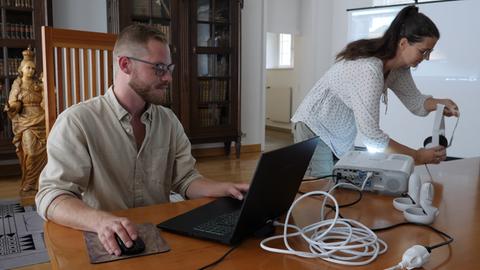 Ein Mann am Schreibtisch, neben ihm greift eine Frau nach einer VR-Brille