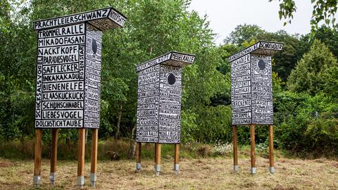 Das Bild zeigt das Kunstwerk Wolkenkuckucksheim des niederländischen Künstlers Fredie Beckmans: drei weiß bemalte Vogelhäuser in der Größe von Hochsitzen für die Jagd, beschrieben mit schwarzen Wörtern.