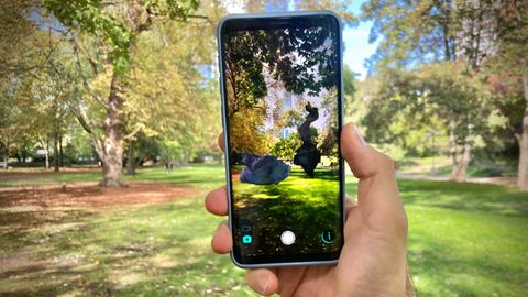 Das Bild zeigt eine Hand, die ein Smartphone hält. Dahinter ist ein Park zu sehen. Auf dem Handy-Bildschirm sieht man den Park, allerdings ergänzt um zwei Kunstobjekte von Florian Adolph.