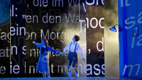 Das Bild zeigt eine Szene aus "Hänsel und Gretel" in der Oper Frankfurt. Zu sehen sind zwei Schauspielerinnen, die Hänsel und Gretel darstellen: links mit zwei langen geflochtenen Zöpfen und einem Kleid, rechts mit Hosenträgern und kurzen Haaren. Sie sind blau beleuchtet. Hinter ihnen sind Worte auf die Wand projiziert.