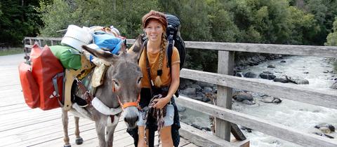 Frau mit Esel und viel Gepäck auf einer Brücke.