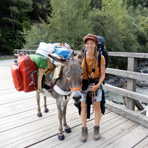 Frau mit Esel und viel Gepäck auf einer Brücke.