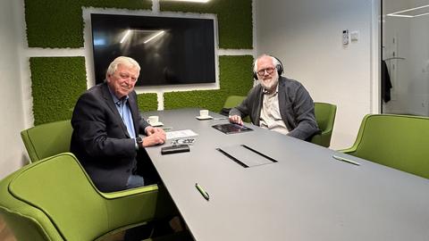Zwei Männer sitzen an einem Tisch, im Hintergrund grüner Wandschmuck