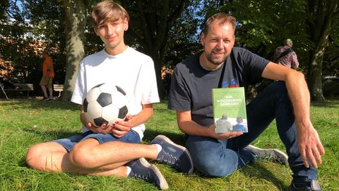 Jason und sein Vater Mirco sitzen auf einer Wiese. jason hält einen ball, sein Vater das Buch "Wir Wochenendrebellen".
