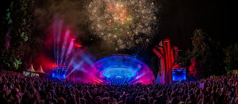 Feuerwerk hinter einer Bühne, im Vordergrund eine große Menge Publikum von hinten, bei Nacht fotografiert