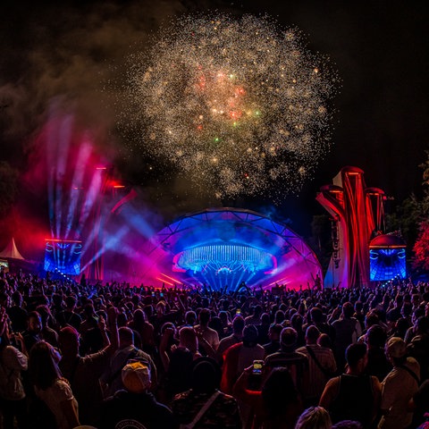 Feuerwerk hinter einer Bühne, im Vordergrund eine große Menge Publikum von hinten, bei Nacht fotografiert