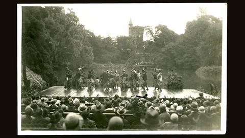 Historisches Foto. In einem Park ist eine Bühne mit tanzenden dunkelhäutigen Menschen mit Lendenschürzen zu sehen. Davor sitzen viele Menschen, die das Publikum bilden.