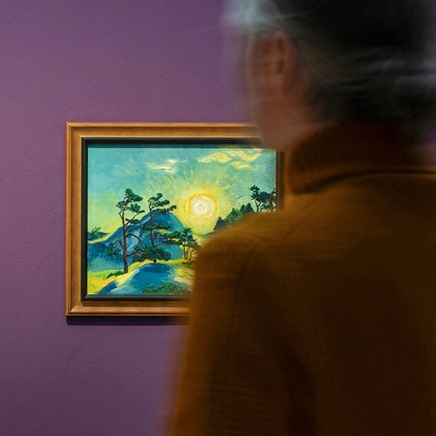 Ein Mensch steht vor zwei gerahmten Gemälden auf einer lilafarbenen Wand