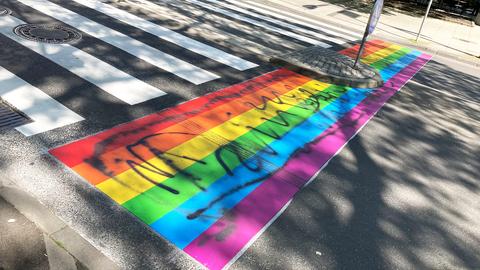 Neben einem klassischen Zebrastreifen mit weißen Querstreifen ein regenbogenfarbener, der so aufgemalt wurde, als würde man über einen Regenbogen gehen. Auf dem Regenbogen sind mit schwarzem Stift unlesbare Zeichen aufgebracht.