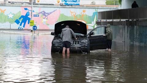 Zwei Menschen stehen wadenhoch im Hochwasser um ihr Auto herum, das vom Wasser umspült ist. 