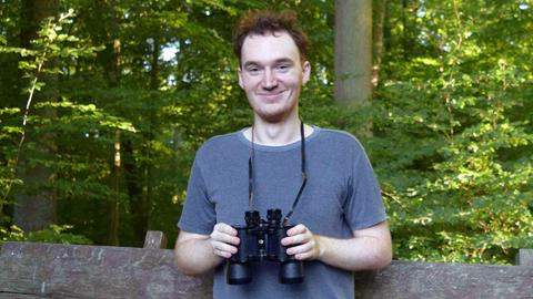 Ein Mann sitzt auf einer Bank im Wald mit einem Fernglas in den Händen und lächelt in die Kamera.