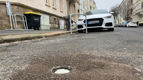 Das Foto zeigt aus der Froschperspektive eine Straße mit parkenden Autos. Im Vordergrund ein Loch im Boden, zur Installtation von Fahrradbügeln, die nebendran uneingebaut an einer Hauswand lehnen. 
