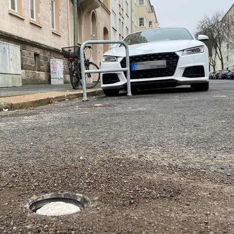 Das Foto zeigt aus der Froschperspektive eine Straße mit parkenden Autos. Im Vordergrund ein Loch im Boden, zur Installtation von Fahrradbügeln, die nebendran uneingebaut an einer Hauswand lehnen. 
