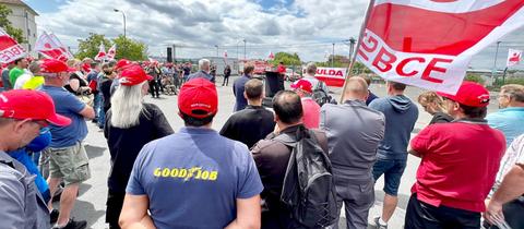 Auf einem Fabrikgelände hören viele Demonstranten mit Fahnen einer Rednerin zu.