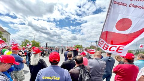 Auf einem Fabrikgelände hören viele Demonstranten mit Fahnen einer Rednerin zu.