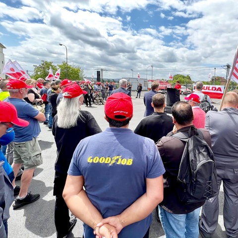 Auf einem Fabrikgelände hören viele Demonstranten mit Fahnen einer Rednerin zu.